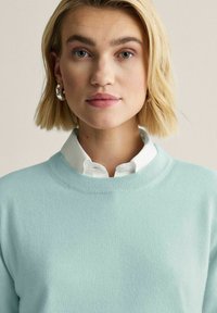 Ljusblå tröja med rund halsringning, bärs över en vit skjorta med krage. Tröjan har en mjuk textur och ribbade ärmslut.