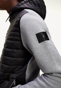 Sort puffervest med en grå sweatshirt som base og et logopatch på ærmet. Har et tekstureret, quiltet design og en hætte.