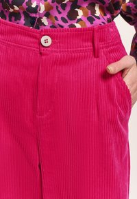 I pantaloni in velluto a coste rosa presentano righe verticali, una chiusura con bottone in vita e tasche laterali. Il tessuto ha una finitura morbida e testurizzata.