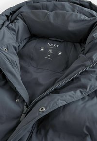 Dunkelgraue Daunenjacke mit durchgehendem Reißverschluss, gestepptem Muster, großem Kragen und einem Innenetikett, das Material- und Pflegehinweise enthält.