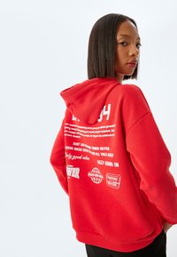 Röd hoodie med vit tryckt text och grafik på baksidan. Materialet verkar mjukt med en avslappnad passform och huva.