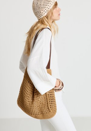 Woman in white outfit wearing a beige crochet hat, carrying a large brown crochet shoulder bag, and wearing bracelets on her wrist.