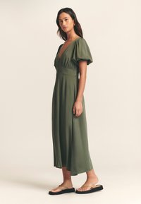 Midi šaty olivově zelené barvy s nafouklými rukávy, hlubokým výstřihem do V a zapínáním na knoflíky vpředu. Modelka v plochých sandálech stojící na neutrálním pozadí.