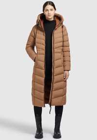 Palton puffer lung, maro, cu glugă, având textură matlasată, fermoar frontal și buzunare laterale. Purtați peste o bluză neagră și pantaloni.