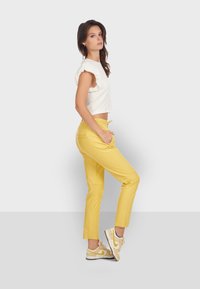 Haut blanc cropped avec des manches volantes, pantalon jaune ajusté et baskets jaunes avec accents blancs, posés sur un fond neutre.