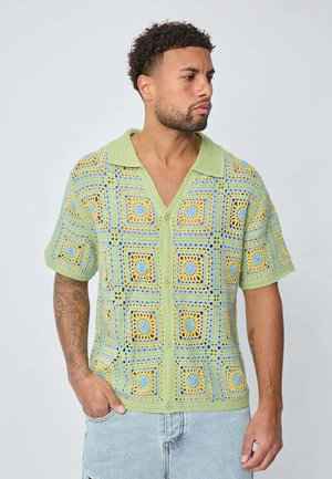 Chemise à manches courtes en crochet de couleur vert clair, présentant des motifs carrés en jaune et bleu, avec un devant boutonné et un col.