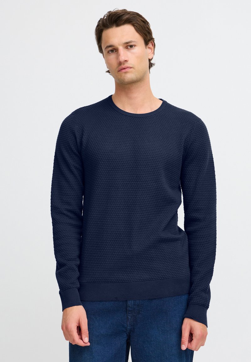 Navy blue textured knit sweater with a round neckline and long sleeves, featuring ribbed cuffs and hem. Worn with blue denim jeans.