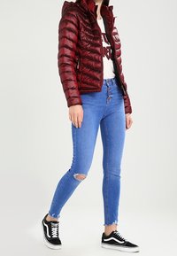 Rød pufferjakke med vandret quiltning, lynlåslukning og hætte. Bæres med lyseblå skinny jeans med huller på knæene og sorte sneakers.