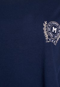 Námořnické bavlněné tričko se dekorativním bílým emblémem s textem a vavřínovými akcenty na levé části hrudi. Hladká textura.