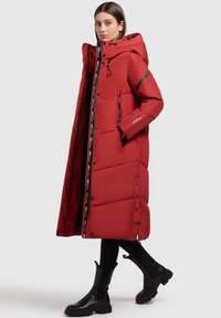 Frau in einem langen roten Puffercoat mit Kapuze, schwarzen Reißverschlussdetails, schwarzen Leggings und schwarzen Stiefeletten vor einem hellen Hintergrund.
