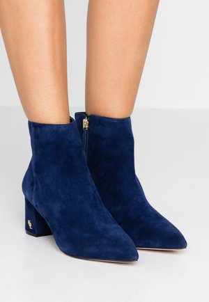 Marineblaue Wildleder-Stiefeletten mit spitzem Zehenbereich, goldenem Reißverschluss und klobigem Absatz mit Logo-Akzent. Glatte Textur, elegantes Design.