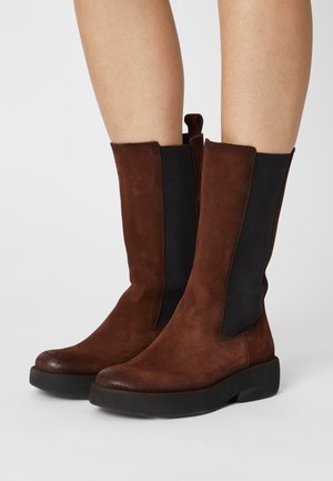 Brune ruskind midt på læggen-støvler med sorte elastiske sidepaneler og chunky sorte såler båret på bare ben mod en ensfarvet baggrund.