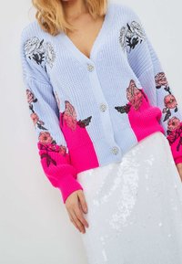 Cardigan lavorato a maglia di colore blu chiaro con accenti rosa e ricami floreali. Presenta grandi bottoni e polsi a coste. Abbinato a una gonna bianca con paillettes.