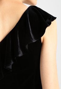 Černý sametový top s jedním volánkovým ramenem. Tkanina má měkkou strukturu s mírným leskem, což umocňuje jeho elegantní vzhled.