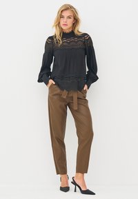 Blouse noire avec détails en dentelle, devant plissé et manches longues volumineuses, associée à un pantalon marron taille haute et des talons noirs.