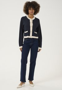 Navy cardigan with cream trim, gold buttons, and two pockets, paired with dark blue jeans and beige heels. Smooth texture and tailored fit.