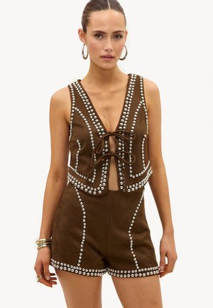 Vrouw draagt een bruine mouwloze vest en bijpassende shorts, beide versierd met zilveren studs, geaccentueerd met creolen en armbanden.