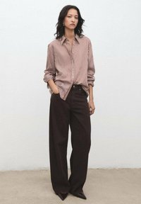 Striped button-up shirt in red and white, paired with wide-leg dark brown trousers. Model stands with hands in pockets against a neutral background.