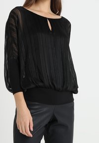 Blusa nera in tessuto trasparente con strisce verticali, collo a occhiello e orlo arricciato. Abbinata a pantaloni di pelle nera.