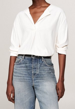 Person, die eine lockere weiße Bluse mit langen Ärmeln und aufgekrempelten Manschetten trägt, in hoch taillierte, ausgebleichte blaue Jeans gesteckt, mit einem schwarzen Gürtel.