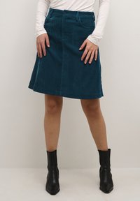 Teal corduroy A-line skirt with a waistline, front pockets, and a smooth texture, paired with black ankle boots.