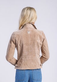 Leren suède jas in tan met een slank ontwerp, geknipte kraag en zilveren hardware. Kenmerken zijn onder andere zijgespen en een logo-opdruk op de achterkant.