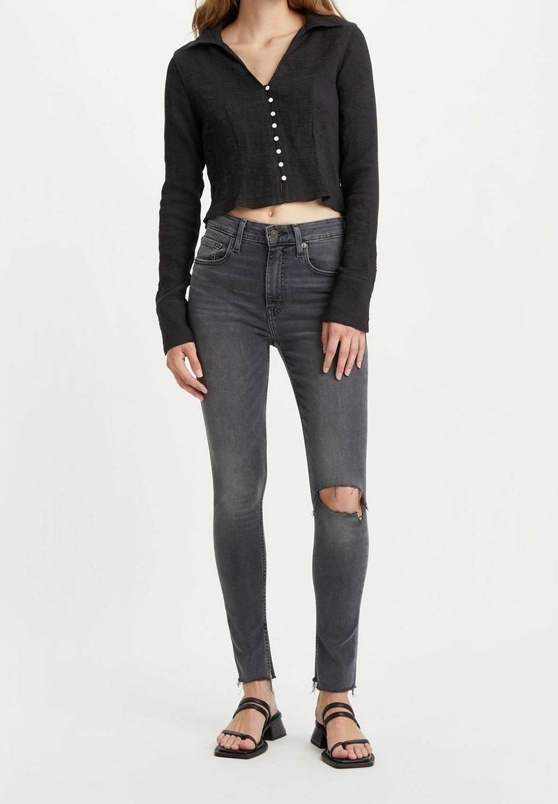 Femme portant un top court noir à manches longues, un jean skinny gris foncé déchiré et des sandales noires à brides, debout devant un fond blanc uni.