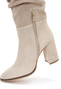 Beżowe zamszowe botki do kostki z okrągłym noskiem, zwężającym się kształtem i 3-calowym słupkowym obcasem; posiadają boczny zamek błyskawiczny oraz delikatnie marszczony górny brzeg.