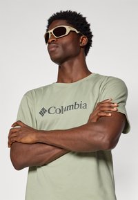 Light green cotton t-shirt with a black "Columbia" logo, worn with tan sunglasses featuring dark lenses, on a person with short, textured hair.