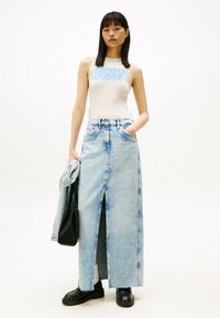 Lichtblauwe denim maxi-rok met een frontale split, gecombineerd met een crèmekleurige tanktop met een blauwe grafische logo. Zwarte schoenen en tas.