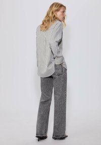 Donna bionda che indossa una camicia a righe larga infilata davanti in jeans dritti grigi effetto acid wash e sandali con tacco neri, in piedi di lato.