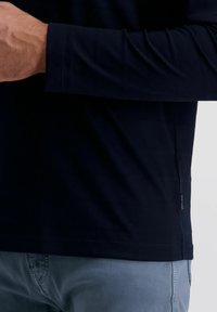 Mann trägt ein langärmeliges schwarzes Shirt und helle blaue Jeans, die Arme über dem Oberkörper verschränkt, mit einem kleinen Etikett am Saum des Shirts.