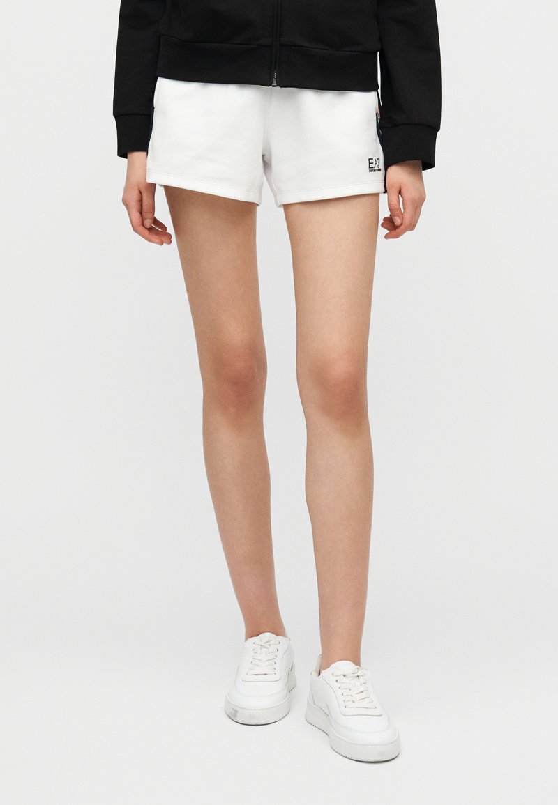 Λευκά αθλητικά σορτς με λεία υφή, διακοσμημένα με μικρό λογότυπο στο πλάι, συνδυασμένα με λευκά αθλητικά παπούτσια και μαύρο μπουφάν με φερμουάρ.