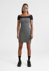 Šedé priliehavé šaty s čiernymi krátkymi rukávmi, vertikálnymi švami a textúrovanou tkaninou; skombinované s čiernymi robustnými teniskami a doplnkami.
