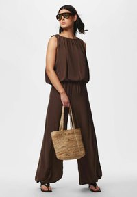 Femme portant une combinaison ample marron sans manches, tenant un sac fourre-tout tressé, de grandes lunettes de soleil et des talons ouverts noirs, debout devant un fond blanc.