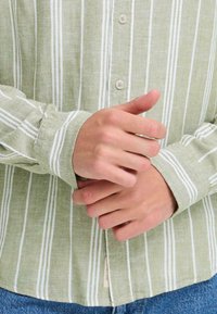 Chemise à rayures vert clair en tissu texturé, avec un devant boutonné et des manches retroussées. Rayures verticales blanches remarquables.