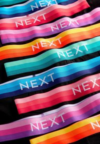 Színes elasztikus szalagok, melyeken a "NEXT" felirat fehér színben szerepel, kék, rózsaszín, piros, lila és narancssárga árnyalataiban megjelenő színátmenetes csíkokkal.