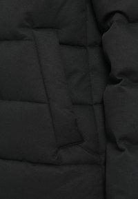 Giacca a vento nera in tessuto imbottito con sezioni trapuntate e una piccola tasca a toppa. La texture appare liscia con una finitura opaca.