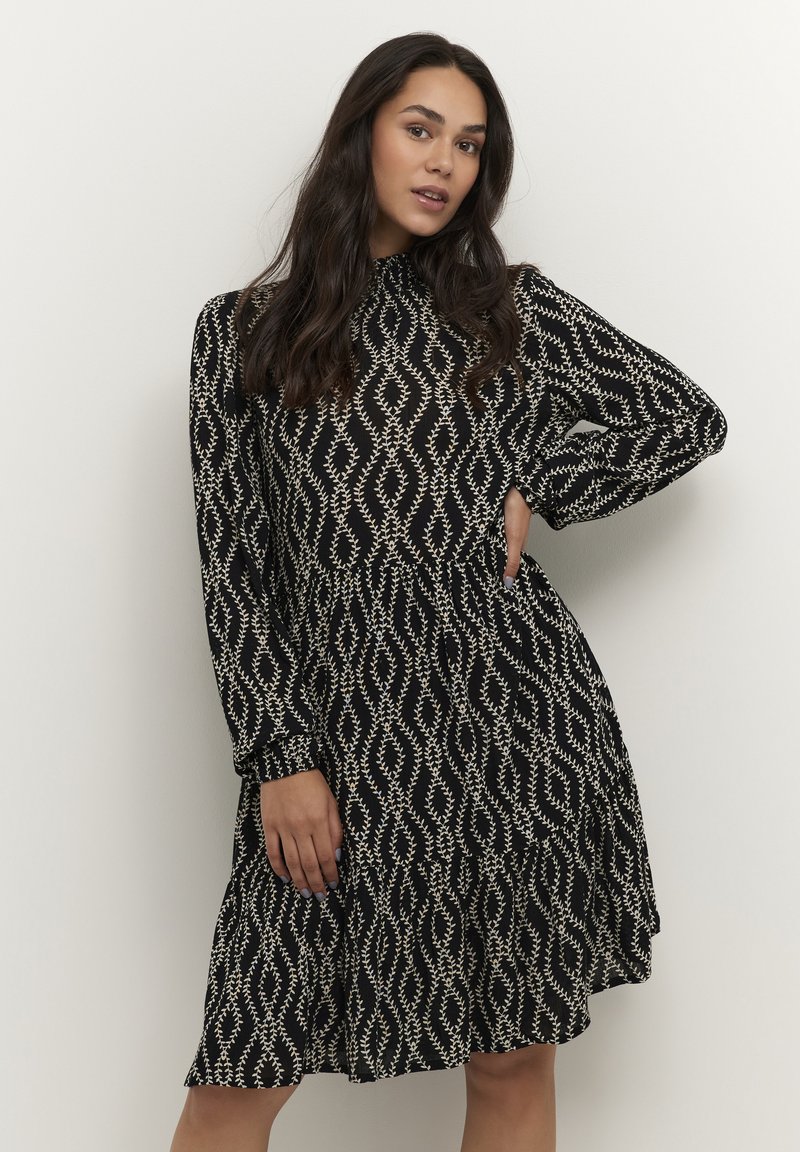Robe noire avec un motif en forme de diamant blanc, dotée de longues manches, d'un col haut et d'une silhouette évasée arrivant au genou.