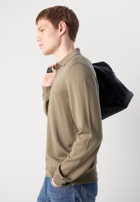 Beige langermet polo-skjorte med ribbet tekstur, to knapper ved kragen, kombinert med en svart ryggsekk med glidelås og håndtak.