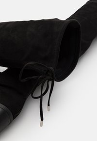 Bota de ante negra hasta la rodilla con tacón de bloque, cremallera lateral y detalle de lazo en la parte superior.