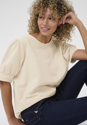 Beige Bluse mit kurzen Puffärmeln und rundem Halsausschnitt. Getragen mit dunkelblauen Jeans, die ein Dreieckselement auf der Gesäßtasche aufweisen.