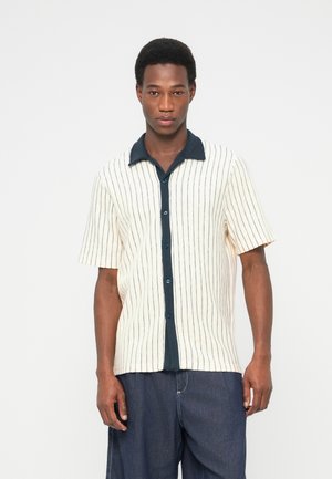 Ung mand med kort krøllet hår iført kortærmet cremefarvet skjorte med sorte lodrette striber og mørkeblå bukser med vide ben på hvid baggrund.