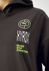 Schwarze Kapuzenjacke mit lebhaften grünen Akzenten, grafischem Text "HYDROX" und "NEVER STOP COMPETING" sowie einem Puma-Logo. Glatte Textur.