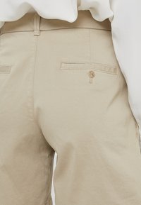 Béžové bavlněné kalhoty mají hladký povrch, dvě zadní kapsy se zapínáním na knoflíky a dvojité švy. Jemná textura zdůrazňuje design.
