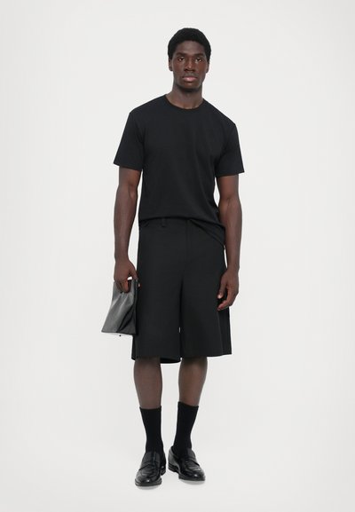 Schwarzes T-Shirt mit kurzen Ärmeln, kombiniert mit schwarzen knielangen Shorts. Halten eine schwarze Clutch, tragen schwarze Socken und Slipper.