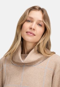 Blonde Frau mit hellen Augen, die einen beigen Pullover mit weitem Kragen und einem dezenten blauen Karomuster vor einem einfarbigen Hintergrund trägt.