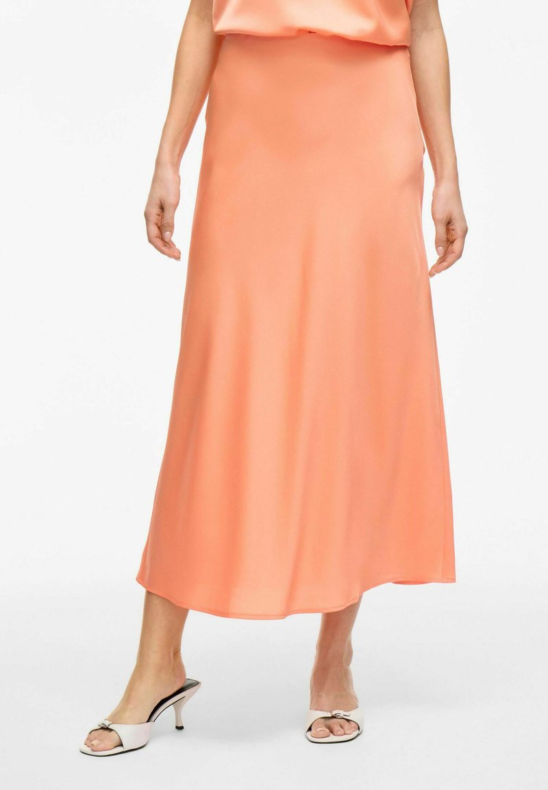 Jupe maxi en satin pêche avec une texture lisse et un léger éclat, présentant une forme A-line et une draperie subtile. Associée à des mules blanches à talons.