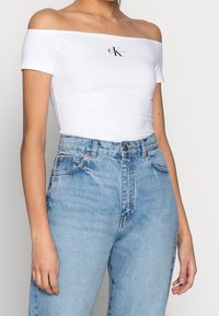 Žena ve bílém off-shoulder topu od Calvina Kleina a v pase vysokých světle modrých džínech na jednobarevném pozadí.