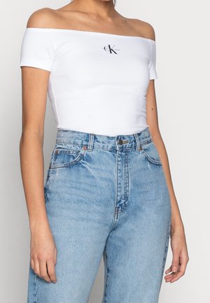 Femme portant un haut Calvin Klein blanc à épaules nues et un jean en denim bleu clair taille haute, sur fond uni.